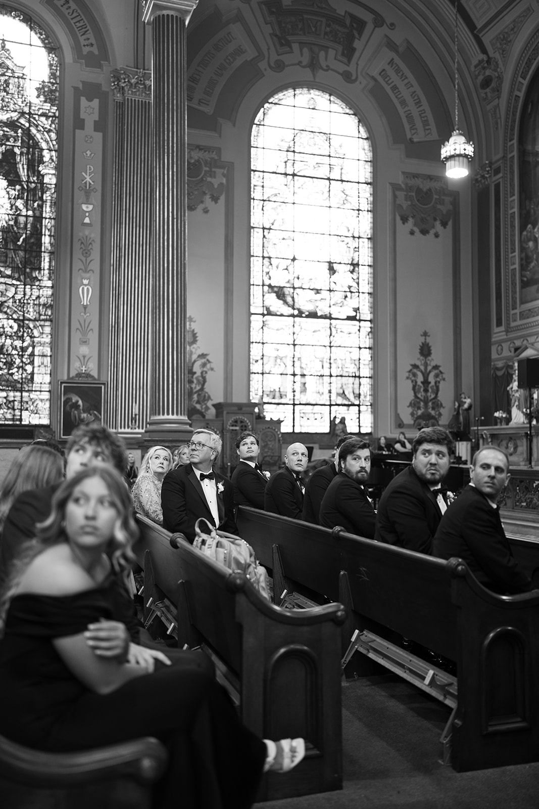 Black-and-white view of wedding guests beneath tall stained-glass windows